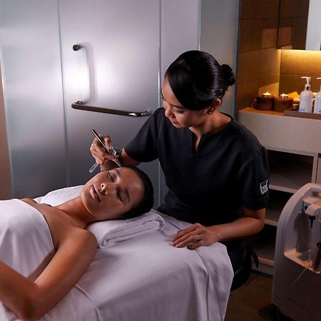 Paradox Singapore Merchant Court At Clarke Quay Екстериор снимка The photo shows a spa or beauty therapy setting where a woman is lying on a treatment table with a towel draped over her. A professional, dressed in a dark uniform, is performing a skincare treatment on her face. Various beauty products are visible o