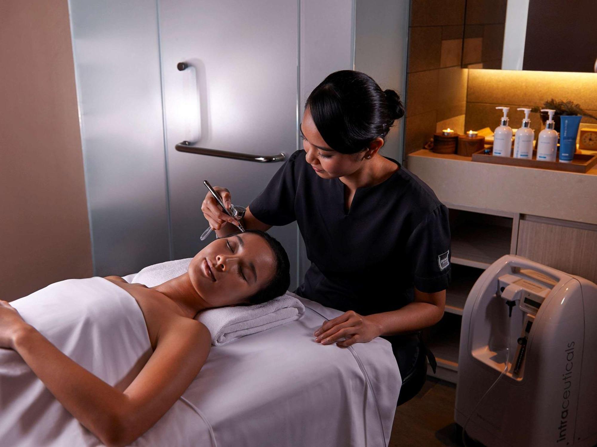 Paradox Singapore Merchant Court At Clarke Quay Екстериор снимка The photo shows a spa or beauty therapy setting where a woman is lying on a treatment table with a towel draped over her. A professional, dressed in a dark uniform, is performing a skincare treatment on her face. Various beauty products are visible o