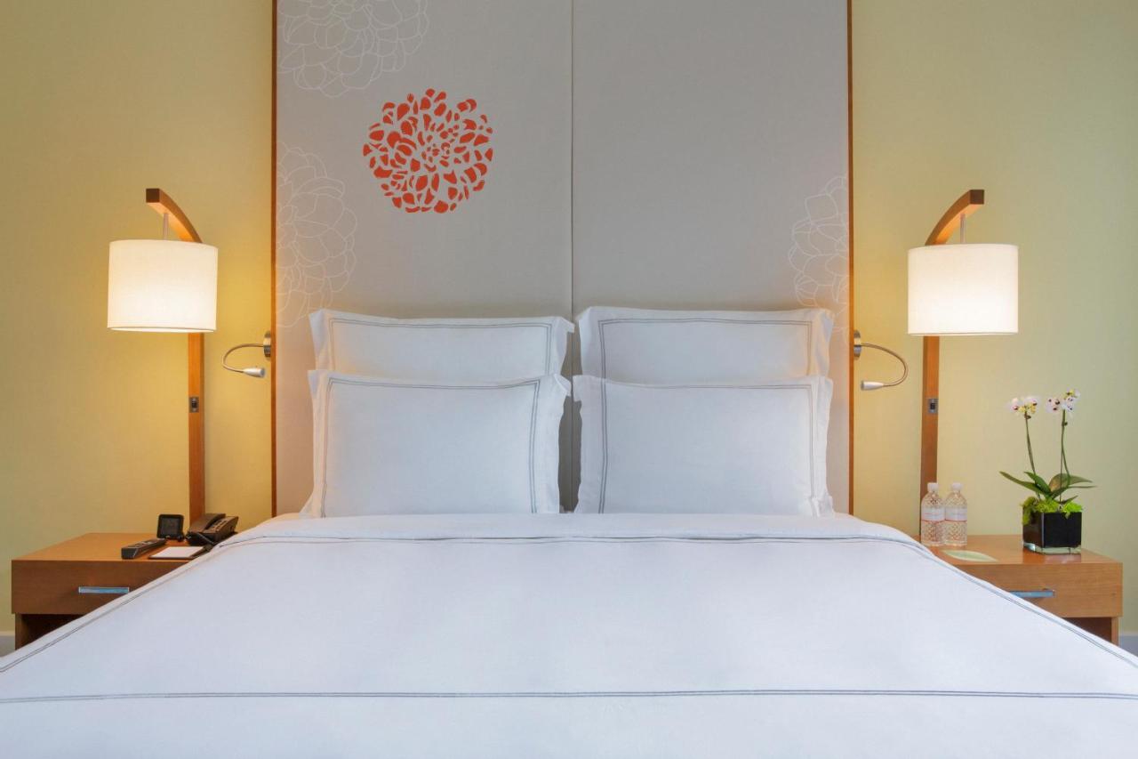 Paradox Singapore Merchant Court At Clarke Quay Екстериор снимка The photo showcases a cozy hotel room with a neatly made bed featuring white linens and two pillows. Behind the bed is a decorative headboard with a simple floral design in orange. On either side of the bed, there are stylish bedside lamps providing 