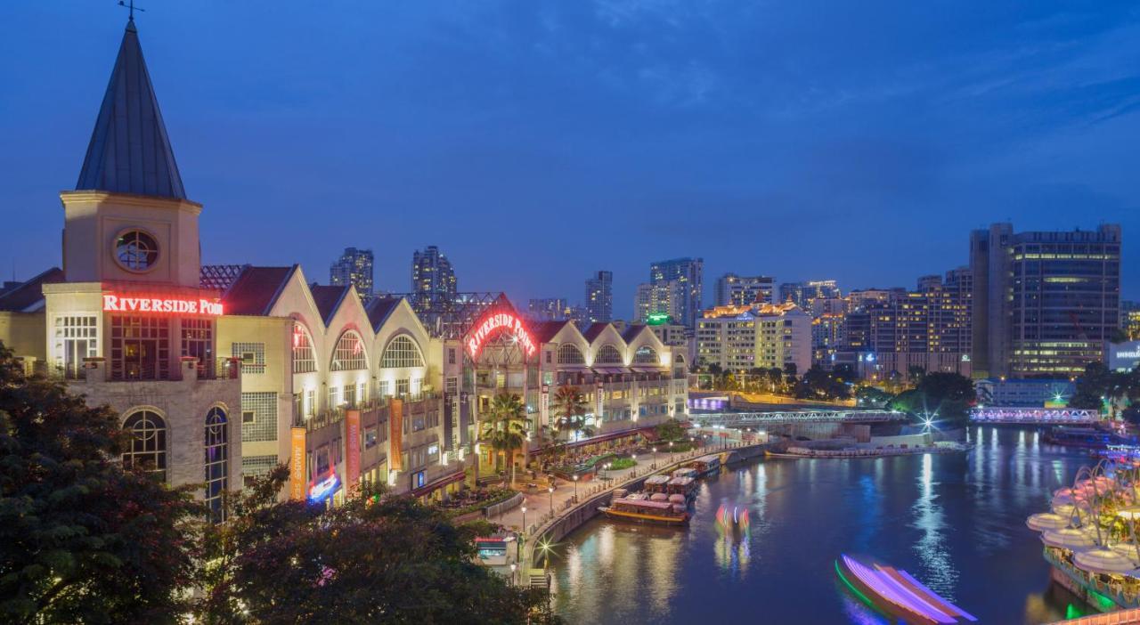 Paradox Singapore Merchant Court At Clarke Quay Екстериор снимка The photo shows a riverside scene at dusk, featuring a waterfront area with modern buildings that have a distinctive architectural style. The structures have pointed roofs and are well-lit, creating a vibrant atmosphere. Along the river, there are bo